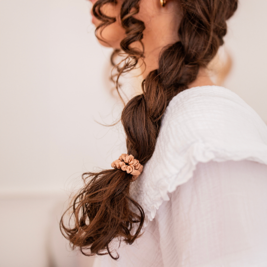 Tresse basse maintenue par un chouchou en soie rose Bachca, sur cheveux bouclés, apportant une finition élégante à une coiffure naturelle.