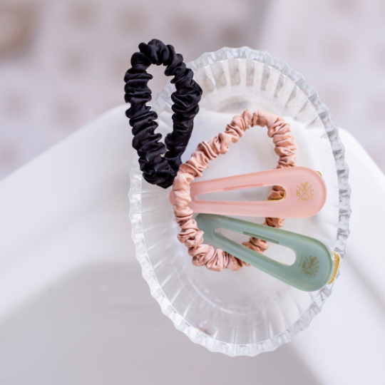 Duo de barrettes Bachca rose dragée et vert d’eau accompagnées de deux chouchous en soie, l’un rose et l’autre noir, posés dans une coupelle en verre transparent sur un lavabo blanc.