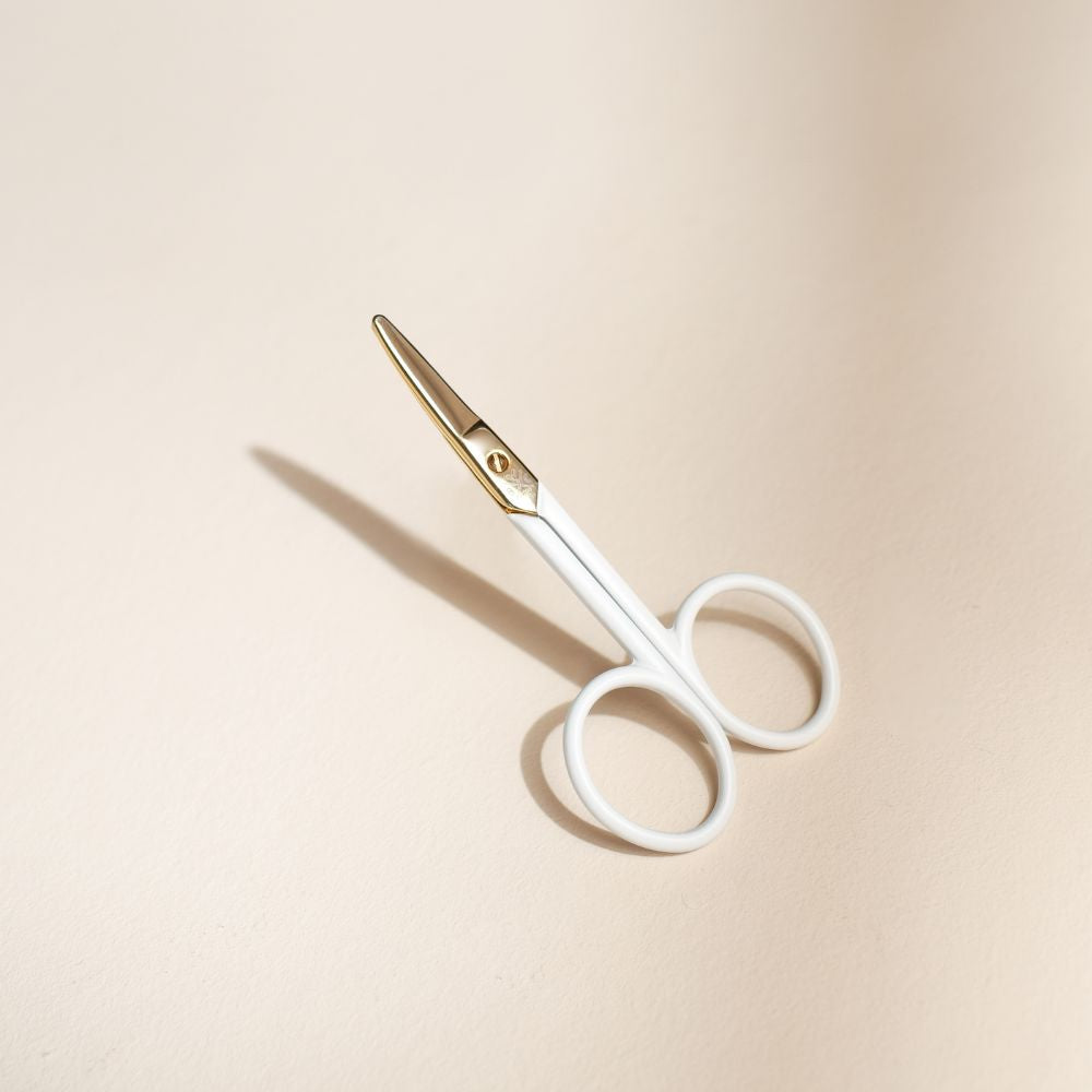 Ciseaux bébé Bachca au design ergonomique, manches blancs et lames dorées arrondies, posés sur un fond beige doux avec ombre portée.