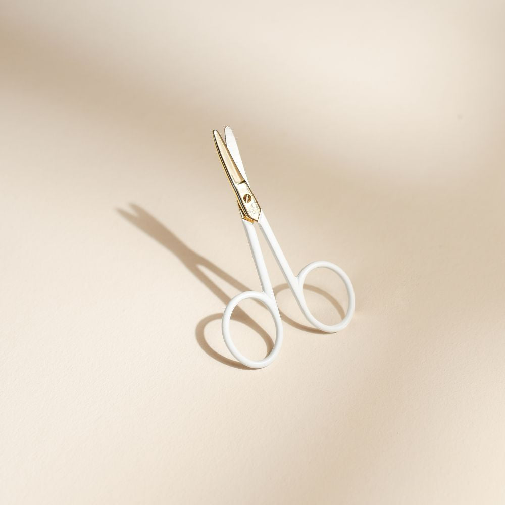 Ciseaux pour ongles bébé Bachca en métal doré et poignées blanches, posés debout sur un fond beige avec une ombre douce.