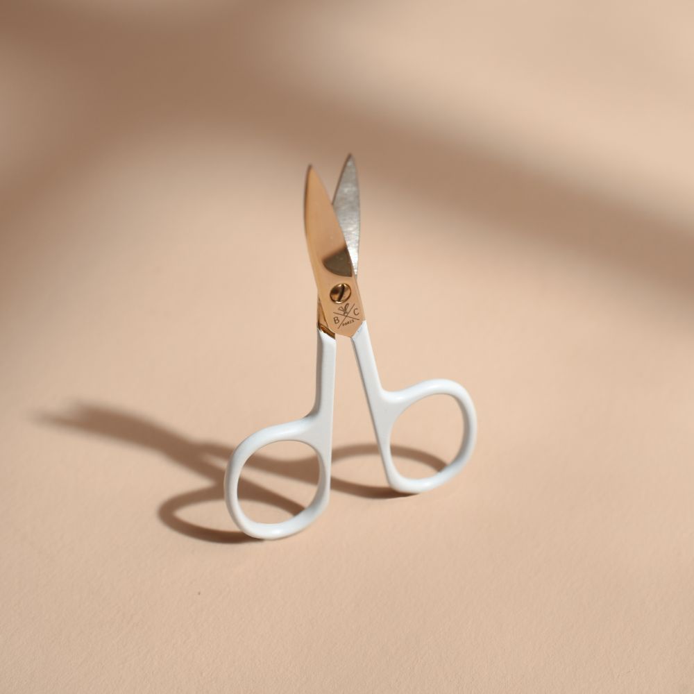 Ciseaux à ongles Bachca avec lames dorées et poignées blanches, photographiés debout sur fond beige avec une ombre marquée.