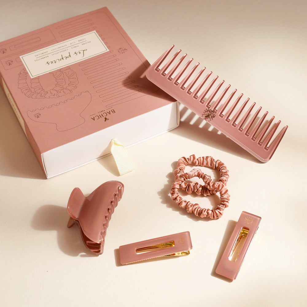 Coffret rose Bachca accompagné d’accessoires assortis disposés sur un fond beige : un peigne large, une pince à cheveux, deux élastiques satinés et deux barrettes rectangulaires dans les mêmes tons poudrés.
