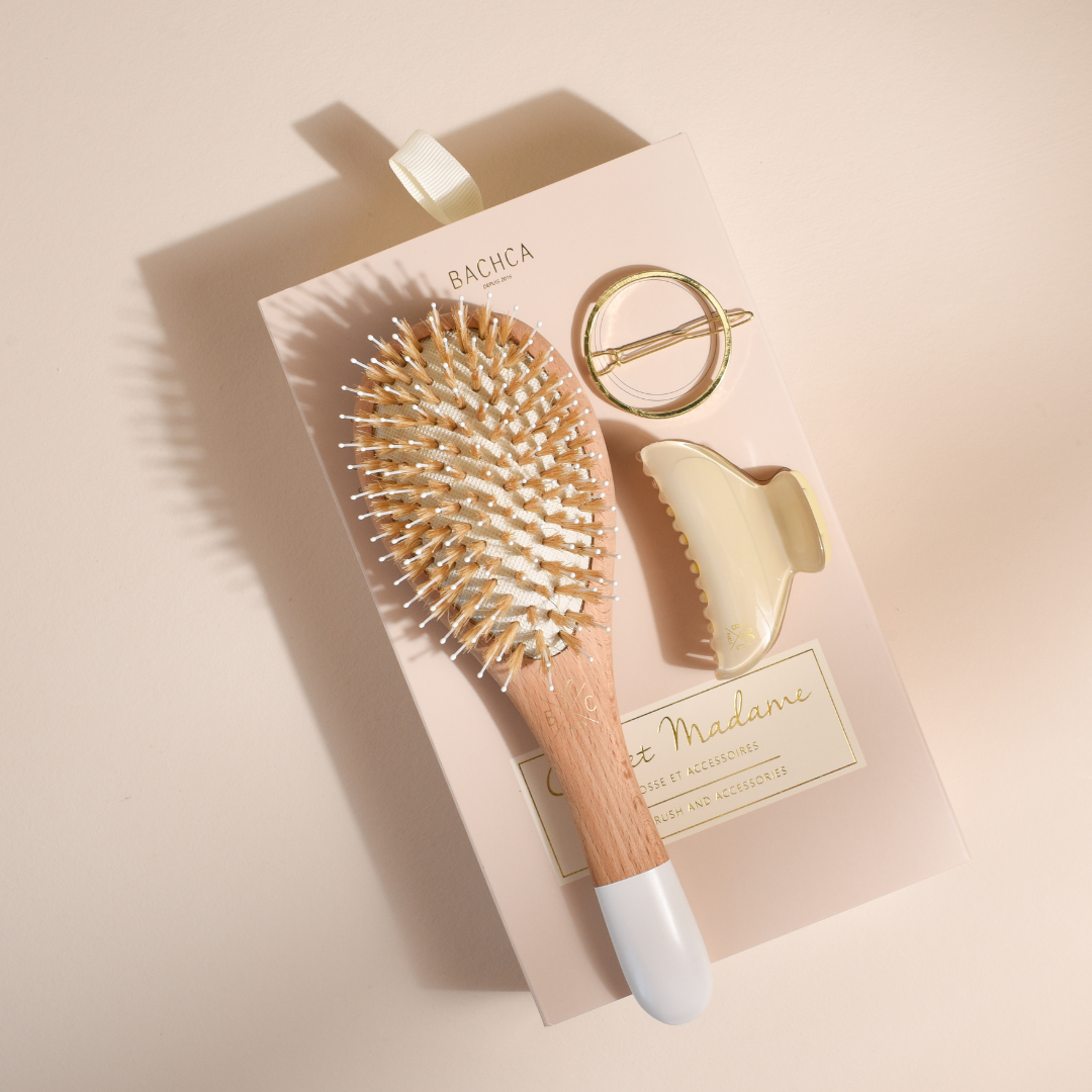 Coffret Madame Bachca présenté à plat, comprenant une brosse à cheveux en bois avec manche blanc, une pince à cheveux crème et une barrette ronde dorée, le tout posé sur un fond beige lumineux.