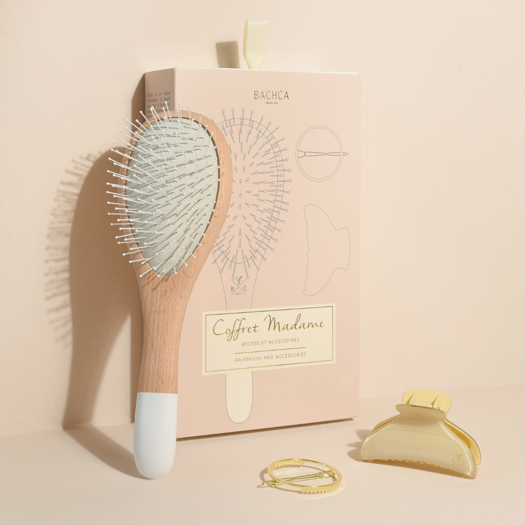 Coffret Madame Bachca en position verticale, accompagné d’une brosse à cheveux picots nylon, d’une pince crème et d’une barrette ronde dorée, sur fond beige poudré.