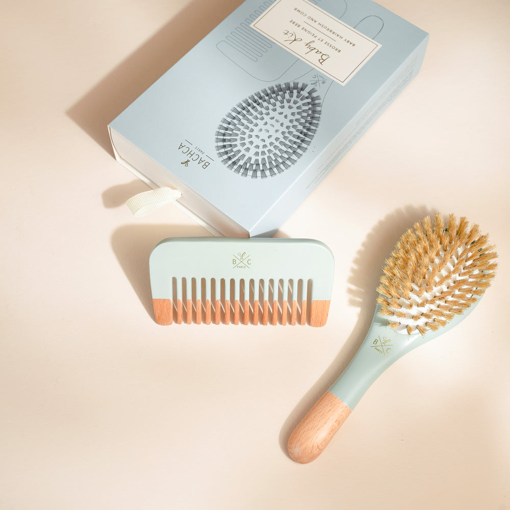 Kit naissance Bachca présenté sur un fond beige clair. À côté de la boîte bleu ciel, on distingue une petite brosse en bois et poils naturels au manche bleu assorti ainsi qu’un peigne coordonné, mêlant bois clair et touches de bleu. La lumière douce projette des ombres délicates sur les accessoires.