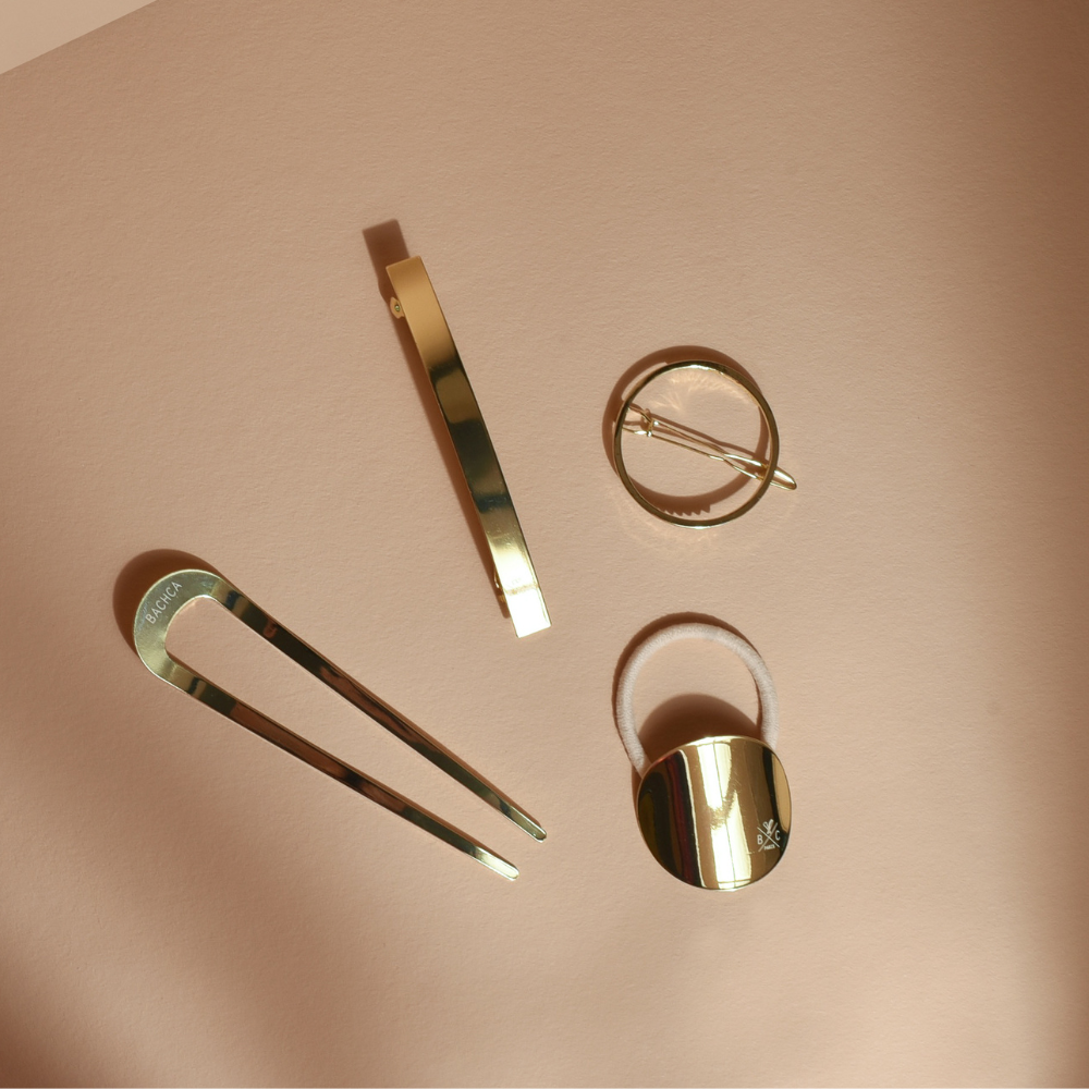 Pic chignon, barrettes et élastique dorés de la marque Bachca posés sur un fond studio rose pâle.