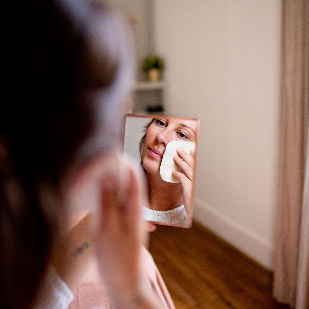 Femme se démaquillant le visage avec un coton lavable Bachca, reflet capturé dans un petit miroir.