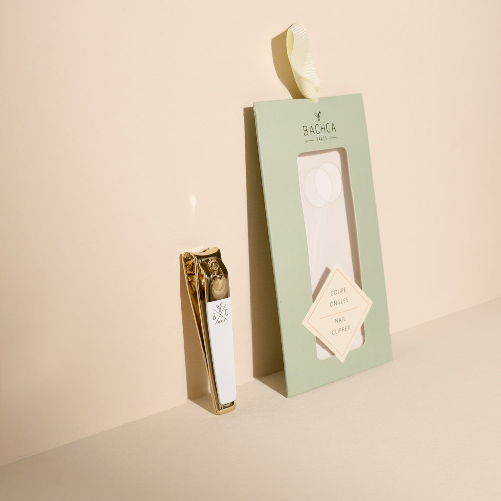 Coupe-ongles doré avec manche blanc siglé Bachca, présenté debout à côté de son étui cartonné vert pastel avec fenêtre transparente et étiquette diamant, sur un fond crème épuré.
