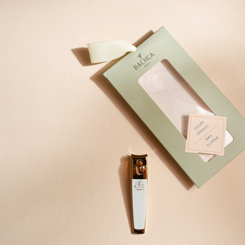 Coupe-ongles doré avec manche blanc siglé Bachca, posé à plat à côté de son étui vert pastel muni d’un ruban crème, sur un fond beige lumineux.