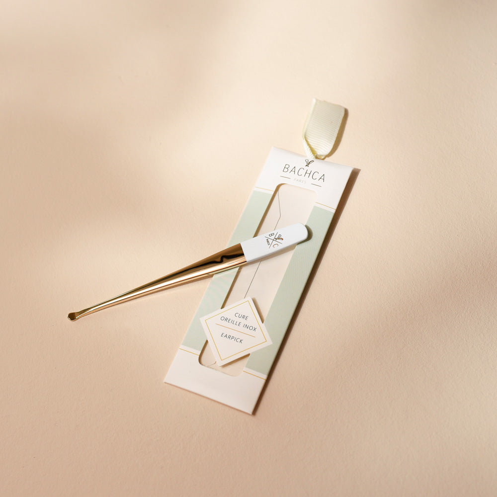 Cure-oreille doré avec manche blanc signé Bachca, présenté dans son étui cartonné aux tons pastel vert et beige, posé sur un fond uni crème.