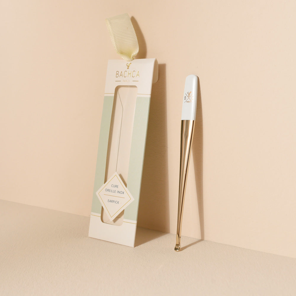 Cure-oreille en métal doré avec manche blanc, posé verticalement à côté de son étui cartonné pastel vert et beige signé Bachca, sur un fond crème minimaliste.