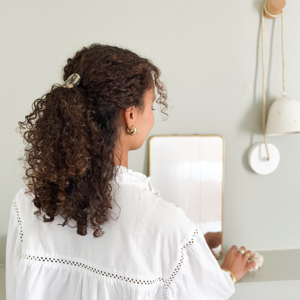 Femme de dos aux cheveux bouclés. Elle porte une demi queue de cheval avec un élastique plaque écaille dans ses cheveux. Les couleurs de la photo sont pastels.