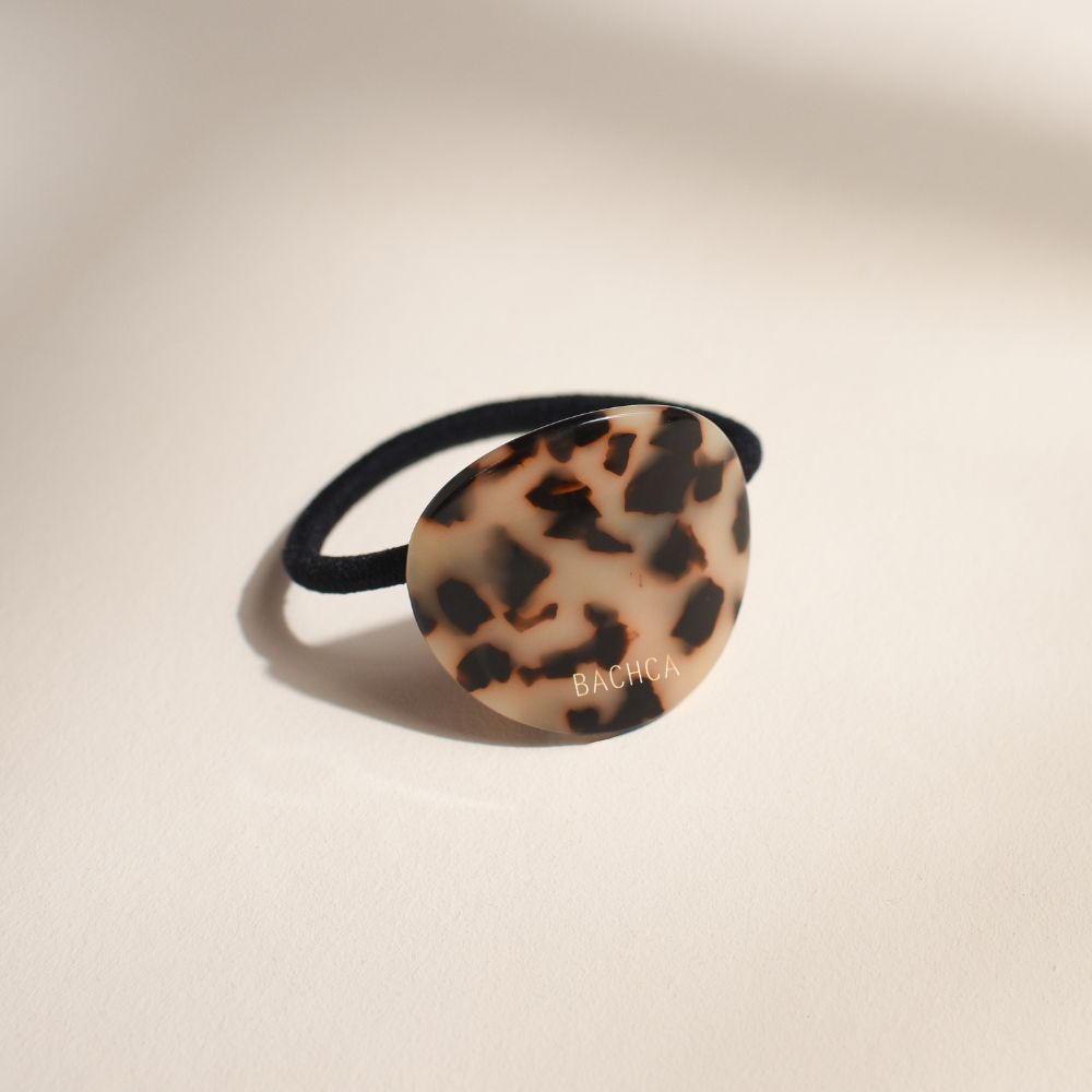 Élastique à cheveux noir avec une plaque ronde écaille de la marque Bachca. Le produit est sur fond rose pâle.