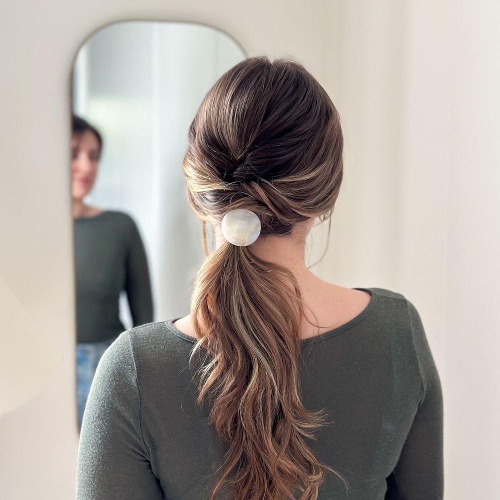 Femme de dos devant un miroir, coiffure élégante avec queue-de-cheval basse maintenue par un élastique plaque nacré Bachca.