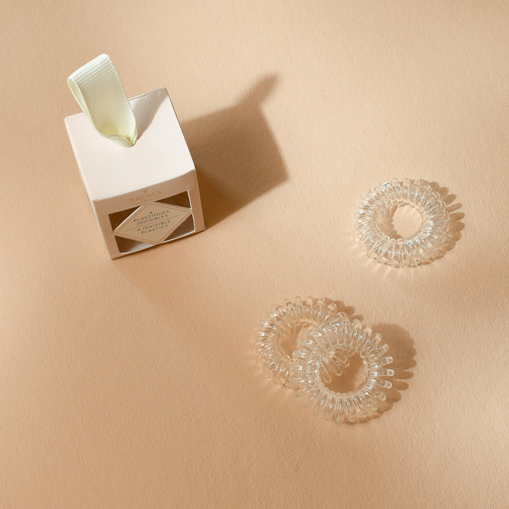Boîte d’élastiques invisibles Bachca avec trois élastiques transparents en spirale disposés à côté, photographiée sur une surface beige.