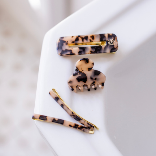 Trois accessoires cheveux Bachca en acétate façon écaille posés sur le bord d’un lavabo blanc : une pince rectangulaire, une petite pince crabe, et deux épingles fines dorées à motif écaille.