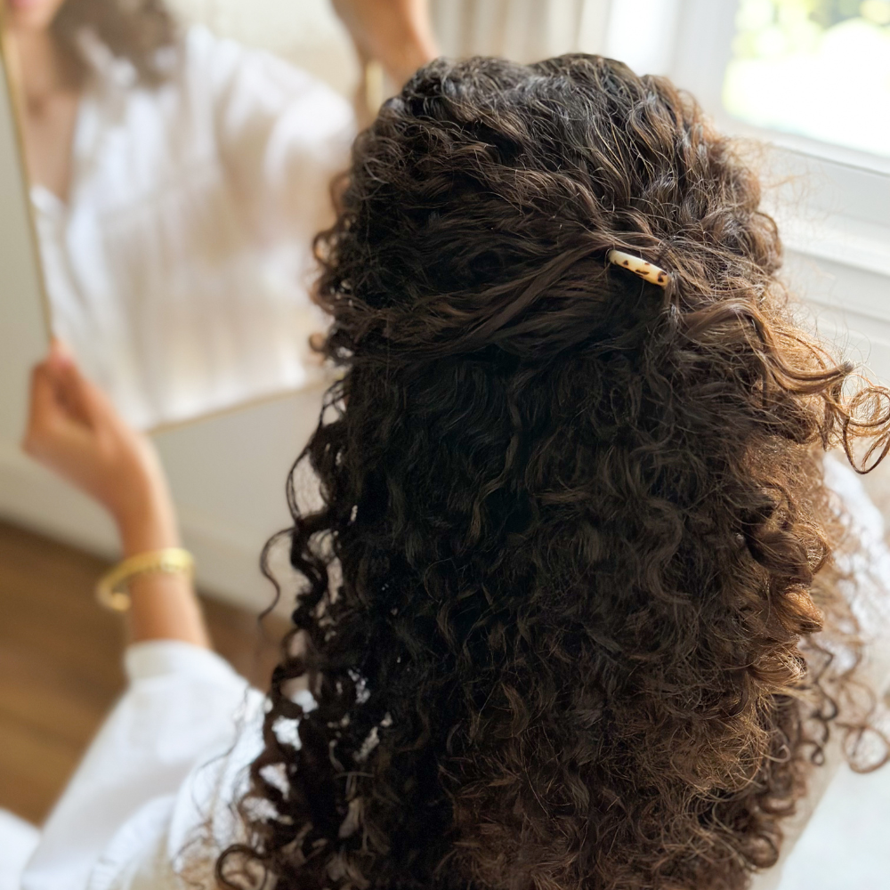 Photo d'une femme aux cheveux bouclés qui porte une épingle à chignon écaille Bachca dans ses cheveux, elle se regarde dans le miroir.