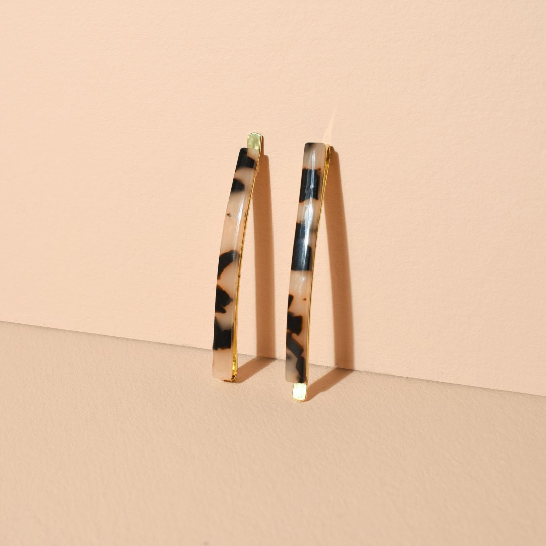 Deux épingles à cheveux fines à motif écaille beige et noir, posées debout contre un mur sur un fond sable uni, mettant en valeur leurs reflets brillants et leur structure dorée.