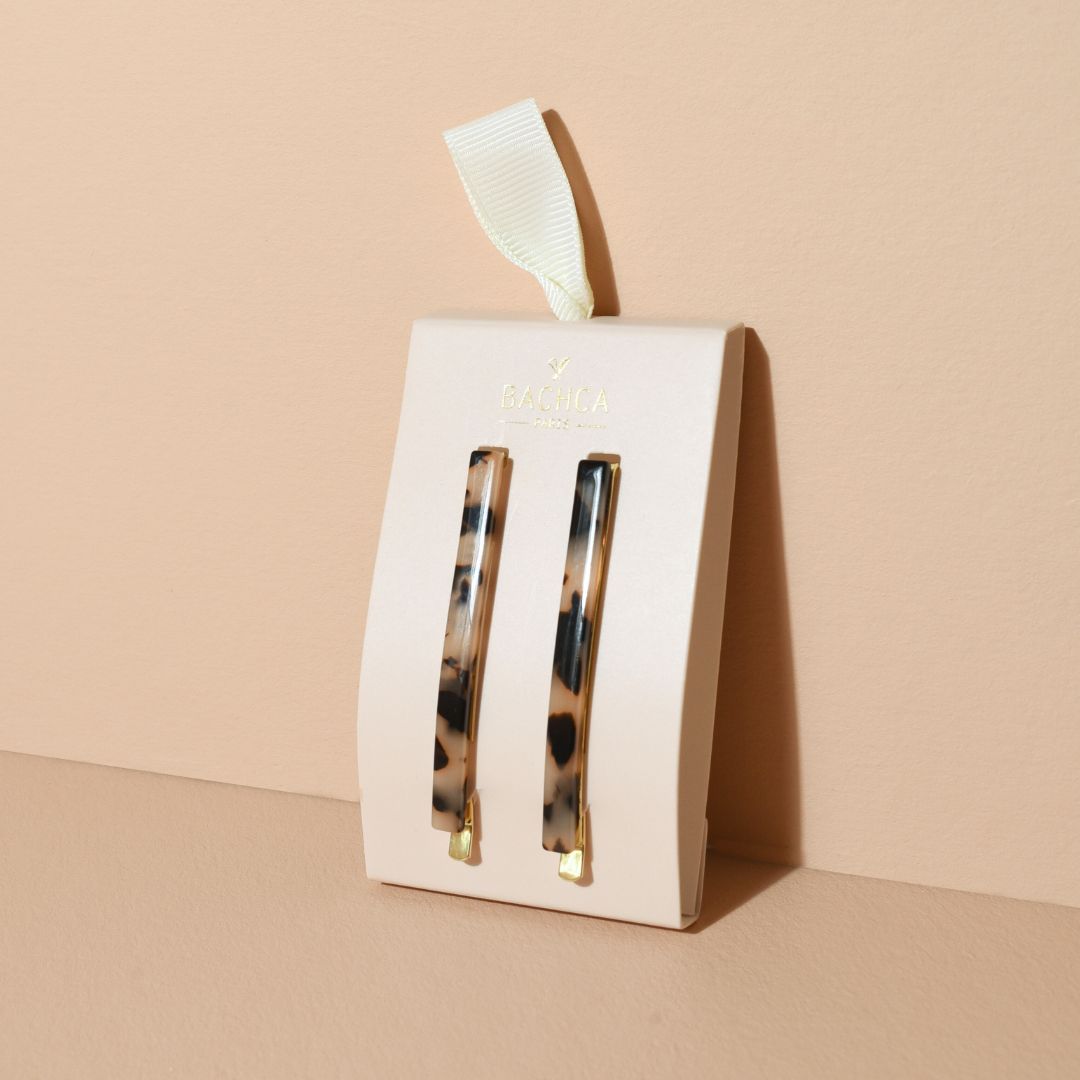Deux épingles à cheveux fines à motif écaille beige et noir, fixées sur leur support en carton crème estampillé du logo doré Bachca Paris, avec un ruban ivoire, posées sur un fond sable chaud.