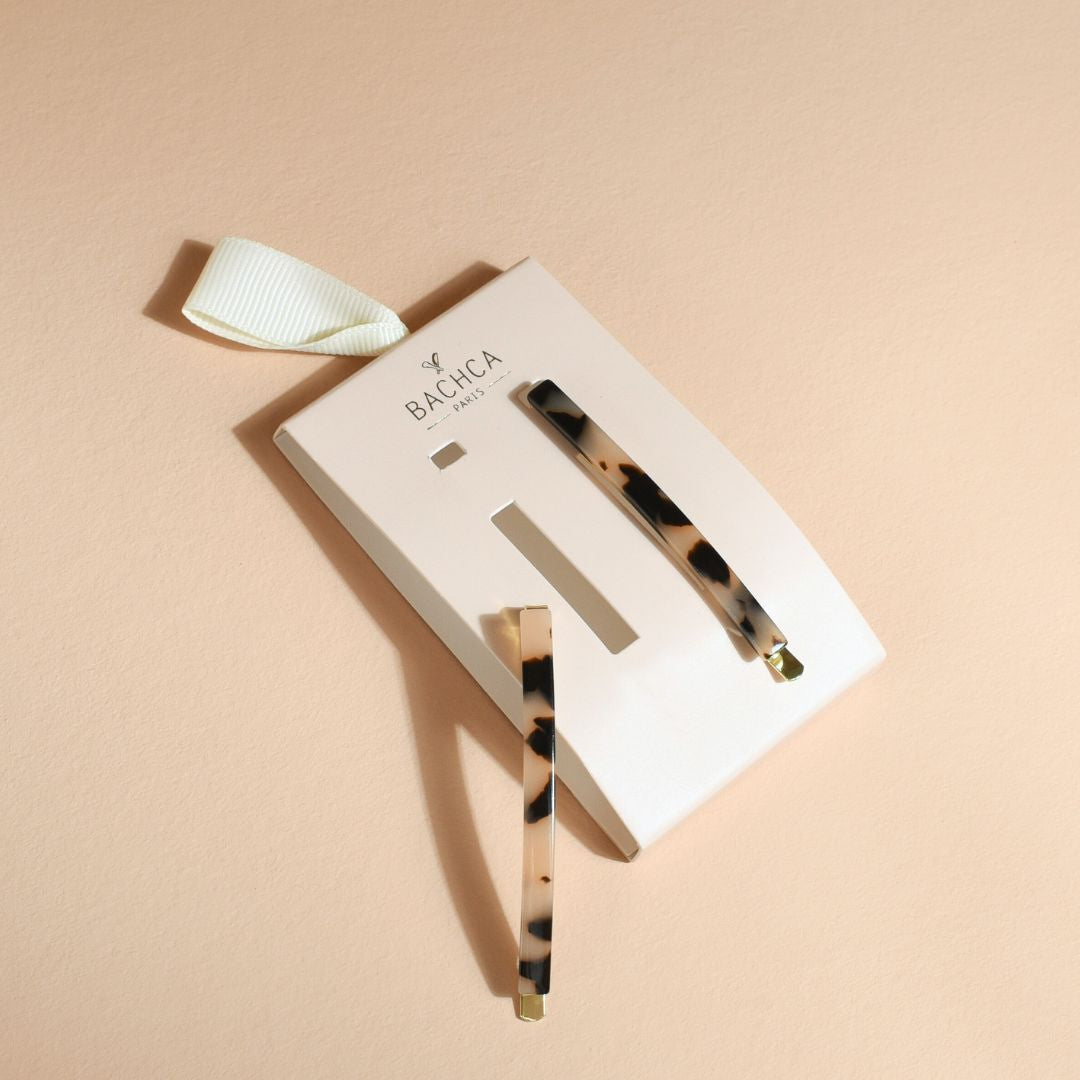 Deux épingles à cheveux fines Bachca à motif écaille beige et noir, dont l’une est détachée et posée à côté du support en carton blanc crème, avec un ruban ivoire, sur un fond sable rosé.