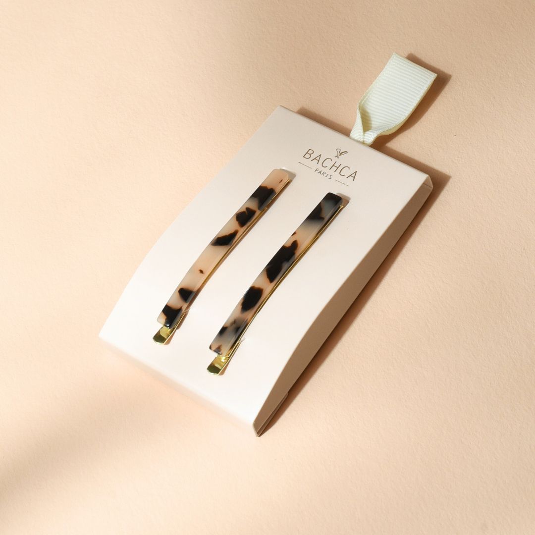 Deux épingles à cheveux fines Bachca à motif écaille beige et noir, présentées sur un support en carton blanc crème estampillé du logo de la marque, posé sur un fond couleur sable rosé.