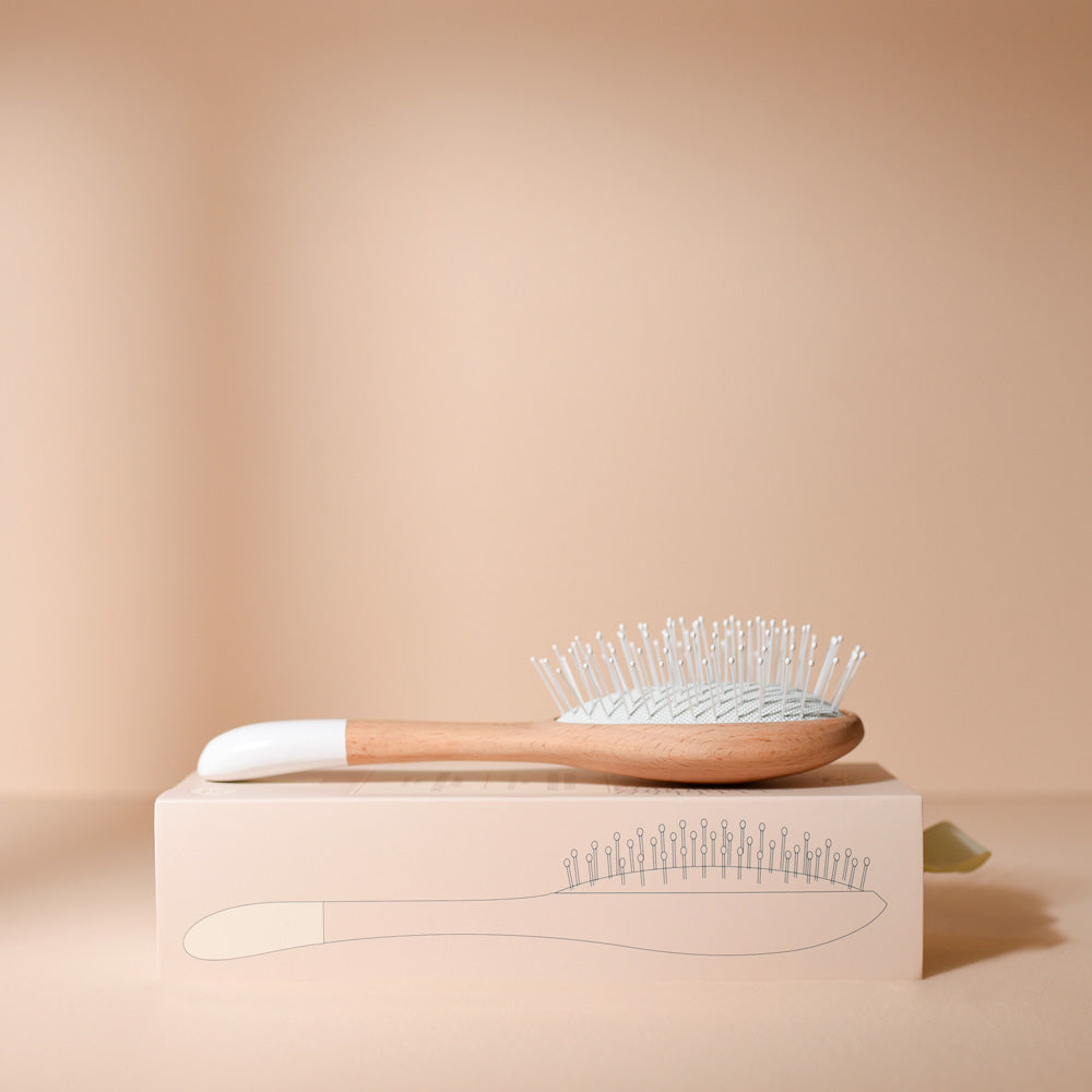 Brosse à cheveux Bachca pour le démêlage et le volume, posée sur sa boîte illustrée, avec manche en bois et picots souples, sur un fond beige élégant.