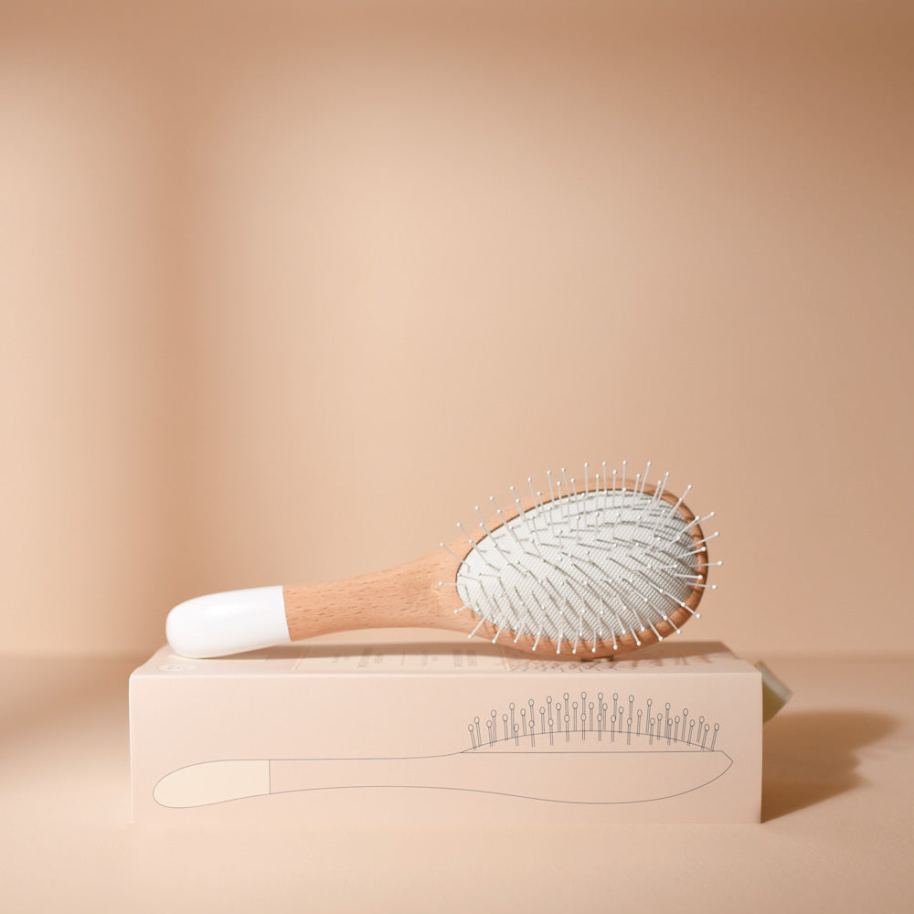 Brosse à cheveux Bachca grand format, spéciale démêlage et volume, posée sur sa boîte illustrée, avec manche en bois naturel et picots souples, sur fond beige.