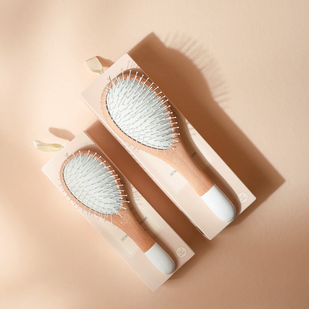 Grande et petite brosses Bachca démêlage et volume, en bois naturel et blanc, posées sur leurs étuis rose poudré.
