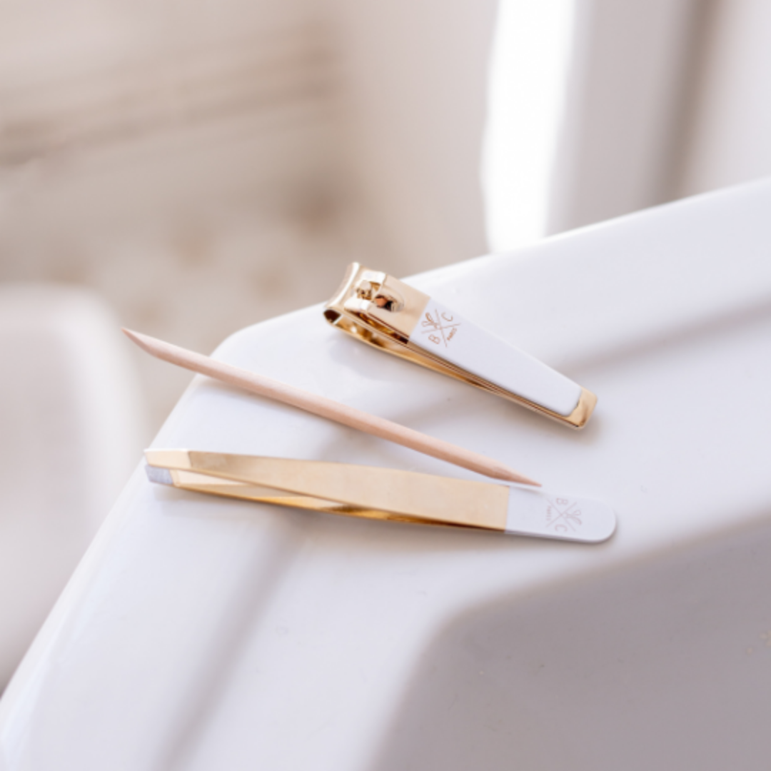 Kit manucure doré et blanc Bachca posé sur un rebord de lavabo : coupe-ongles, pince à épiler et bâtonnet en bois.