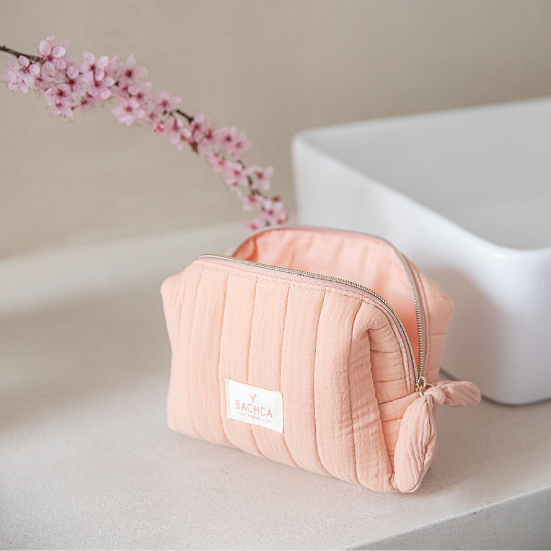 Trousse de maquillage BACHCA en coton matelassé rose, posée près d’un lavabo et d’une branche fleurie.