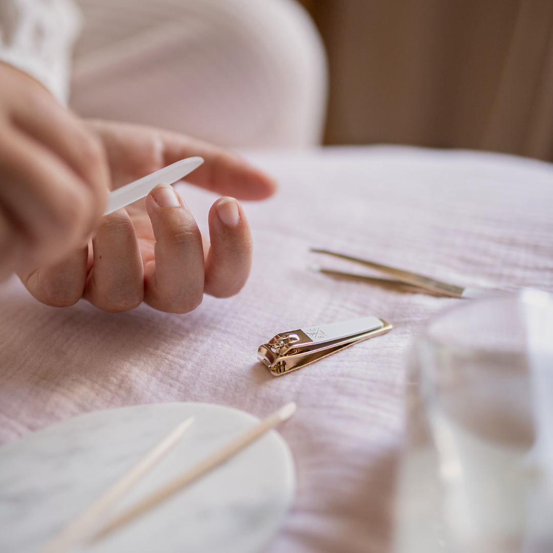 Rituel manucure avec lime à ongles en verre Bachca, coupe-ongles doré et pince à épiler sur fond rose doux.