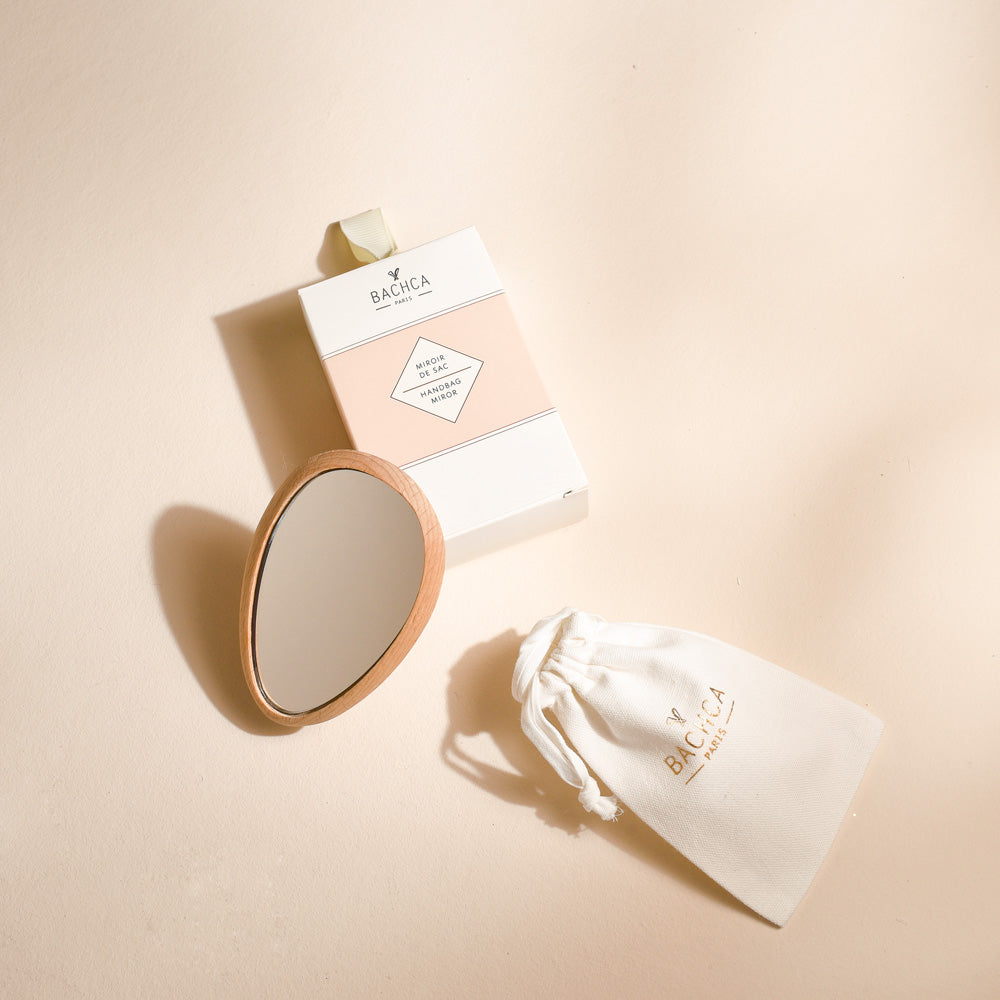 Miroir de poche Bachca en bois clair, de forme goutte, présenté avec son pochon en coton blanc logoté et sa boîte cartonnée rose poudré et blanche, posés sur un fond beige.