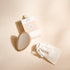 Miroir de poche Bachca en bois clair, de forme goutte, présenté avec son pochon en coton blanc logoté et sa boîte cartonnée rose poudré et blanche, posés sur un fond beige.