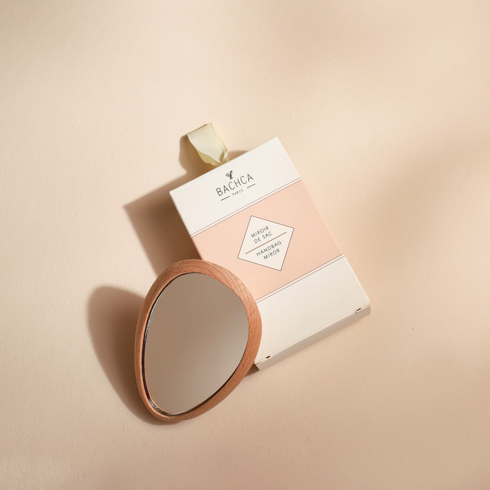 Miroir de sac Bachca en bois clair, de forme goutte, posé à côté de son étui cartonné blanc et rose poudré avec ruban beige, sur un fond uni couleur crème.