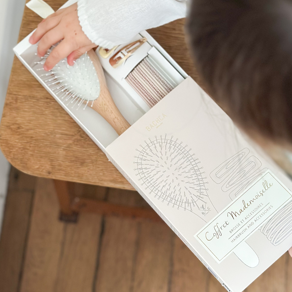 Coffret Mademoiselle de la marque Bachca ouvert par une petite fille, on peut voir une brosse à cheveux, des élastiques fins pastel ainsi que des petites barrettes clic-clac.