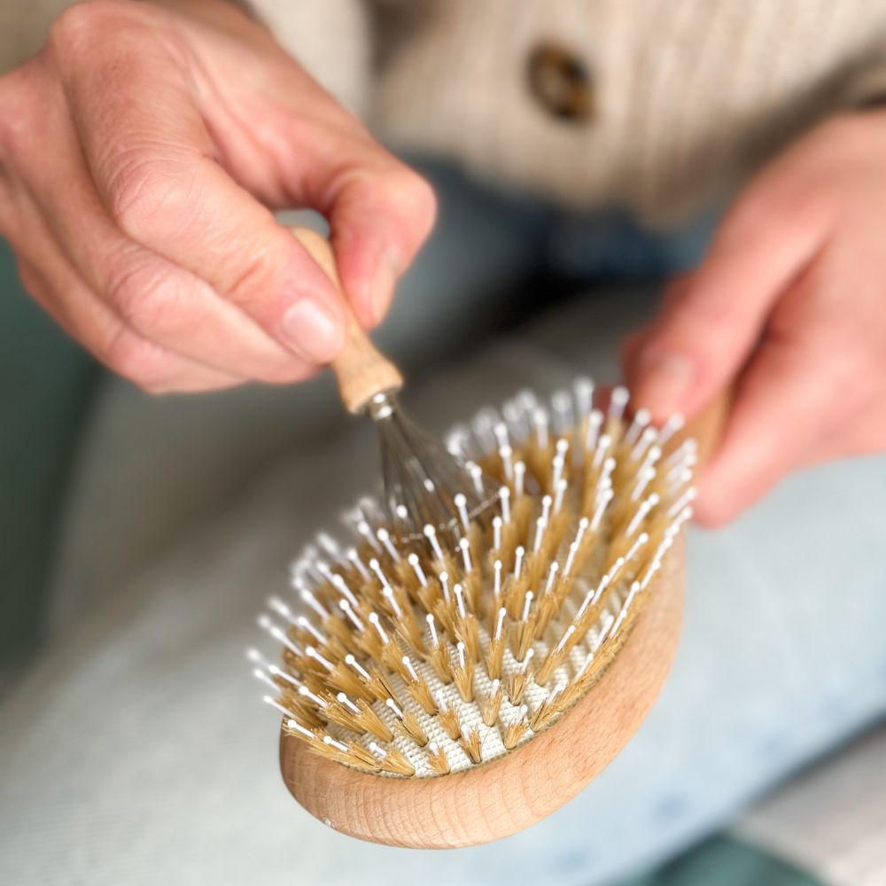 Main nettoyant une brosse à cheveux Bachca en bois avec un peigne métallique pour enlever les résidus.