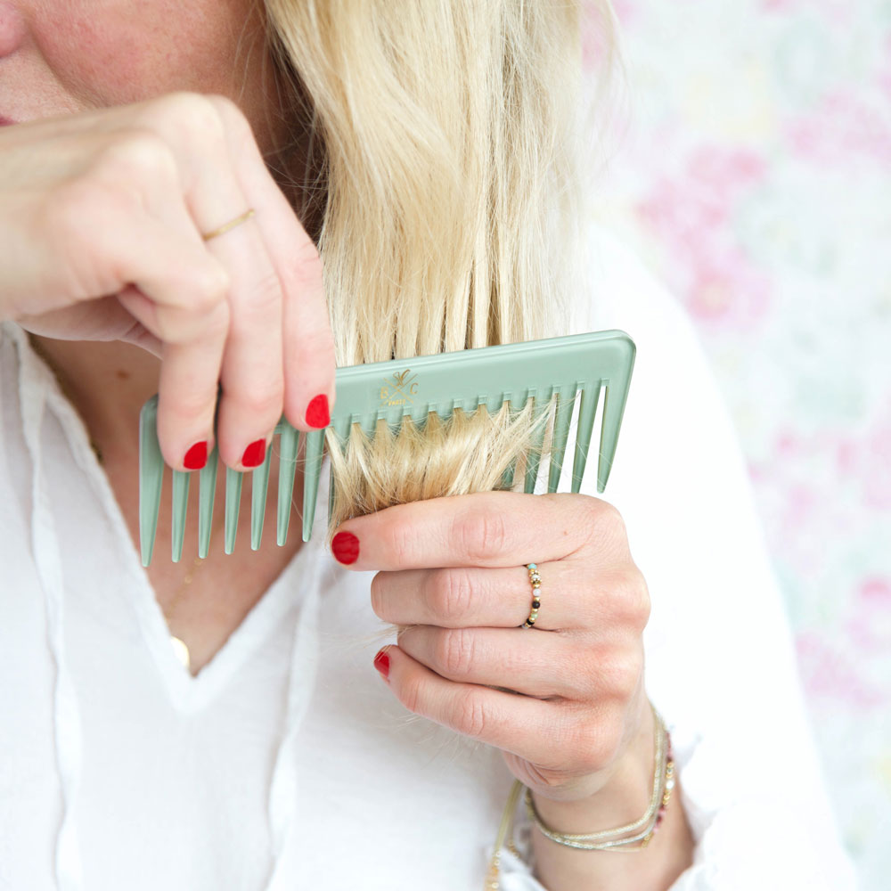 Femme coiffant ses cheveux blonds avec le peigne Opaline vert Bachca, tenue blanche et vernis rouge sur fond floral pastel.