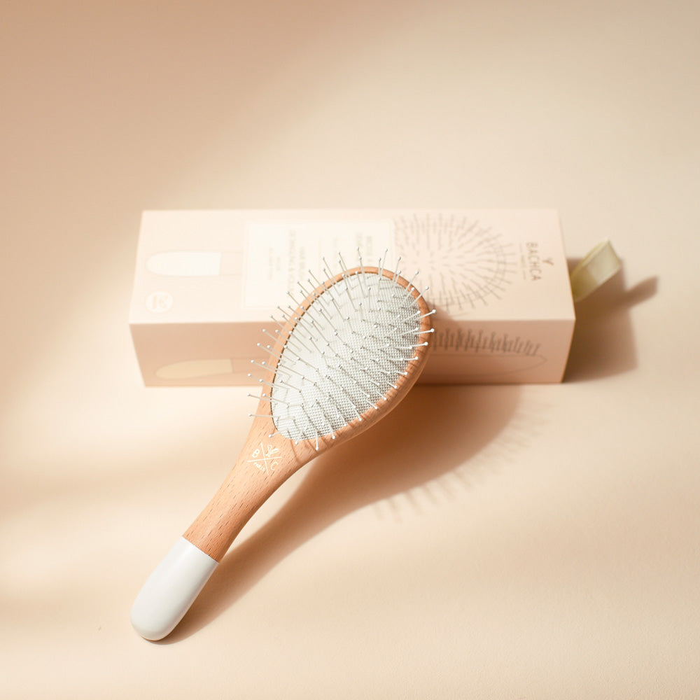 Petite brosse Bachca en bois naturel et blanc, picots nylon argentés, posée devant son coffret beige rosé.