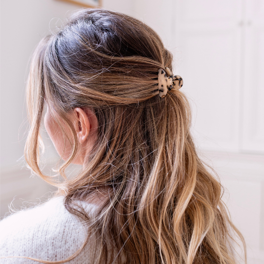Femme aux cheveux blonds foncés et ondulés, coiffée en demi-queue maintenue par une petite pince à motif écaille beige et noir, dans une pièce lumineuse à l’ambiance douce et épurée.