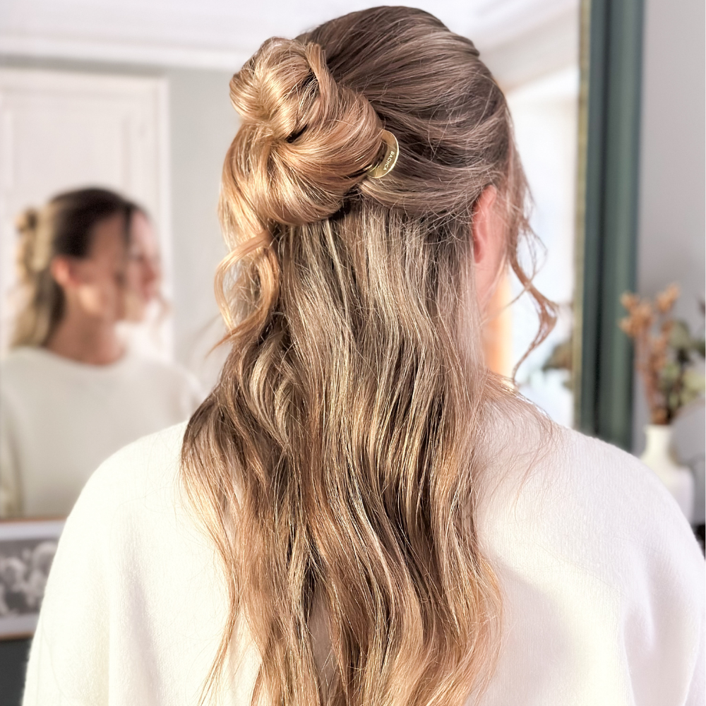Photo d'une femme de dos avec de longs cheveux blonds, elle porte dans ses cheveux un pic chignon doré de la marque Bachca, on voit son reflet dans un miroir en face d'elle.