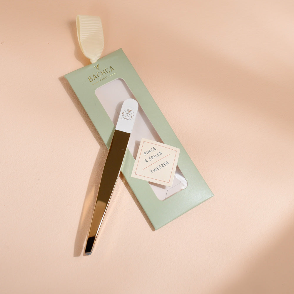 Pince à épiler Bachca dorée et blanche, présentée sur son emballage vert pastel avec étiquette, photographiée sur une surface beige.