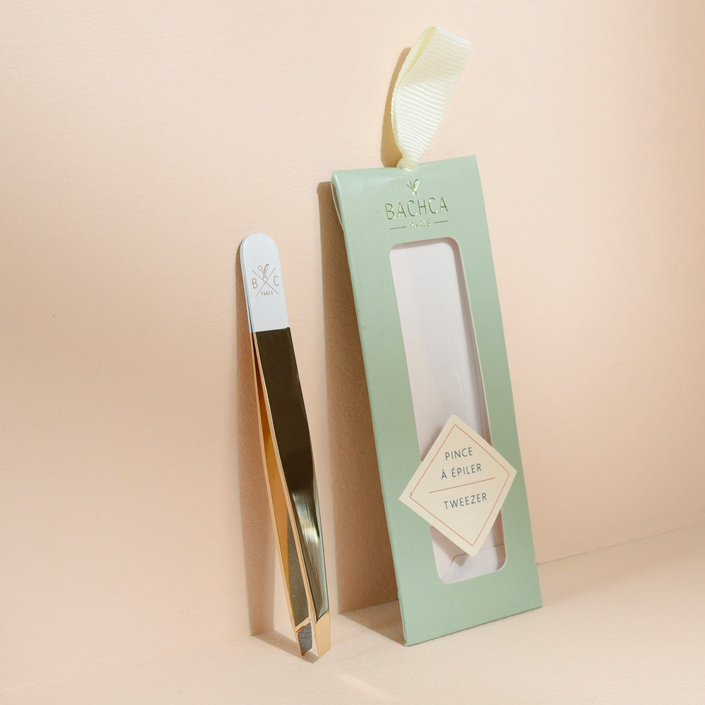 Pince à épiler Bachca dorée et blanche, présentée avec son étui vert pastel, idéale pour une épilation précise et soignée.