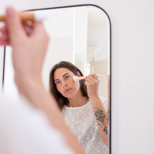 Femme se maquillant devant un miroir à l’aide du pinceau blush Bachca, reconnaissable à son manche en bois naturel et son extrémité blanche, dans une salle de bain à la décoration épurée.