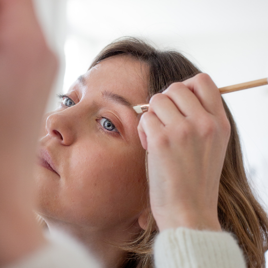 Femme maquillant ses sourcils avec avec un pinceau yeux biseautés Bachca en bois, dans une salle de bain lumineuse au style naturel.
