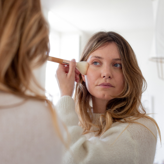 Femme appliquant du blush avec un pinceau poudre Bachca en bois, devant un miroir, dans une salle de bain lumineuse et épurée.