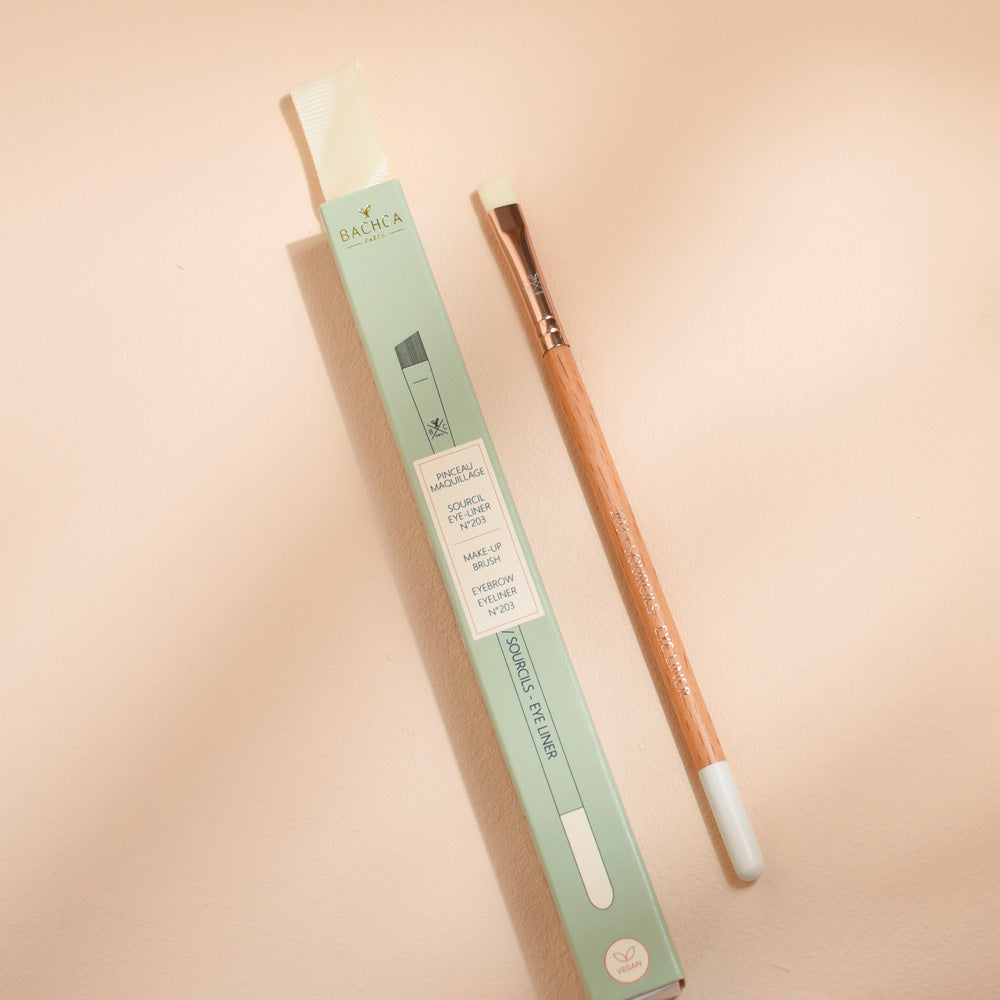 Pinceau sourcils et eye-liner Bachca avec manche en bois et embout biseauté, présenté à côté de son étui vert pastel, photographié sur une surface beige.