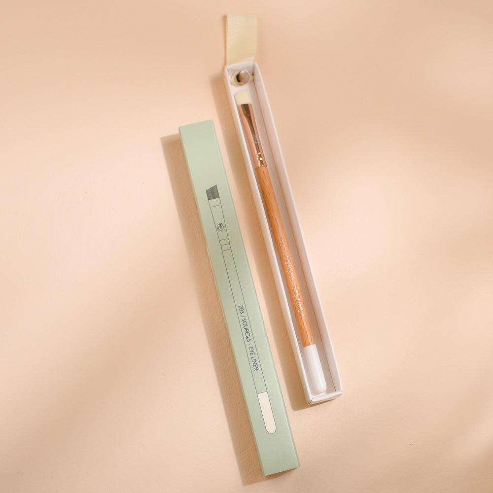 Pinceau sourcils et eye-liner Bachca avec manche en bois et embout biseauté, présenté dans son étui ouvert à côté de son emballage vert pastel, photographié sur une surface beige.