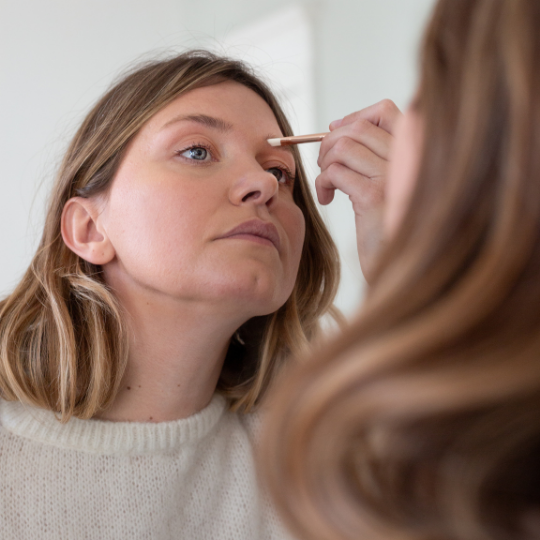 Femme appliquant du fard à paupières avec un pinceau fin Bachca devant un miroir, concentrée sur son maquillage des yeux, vêtue d’un pull blanc en maille.