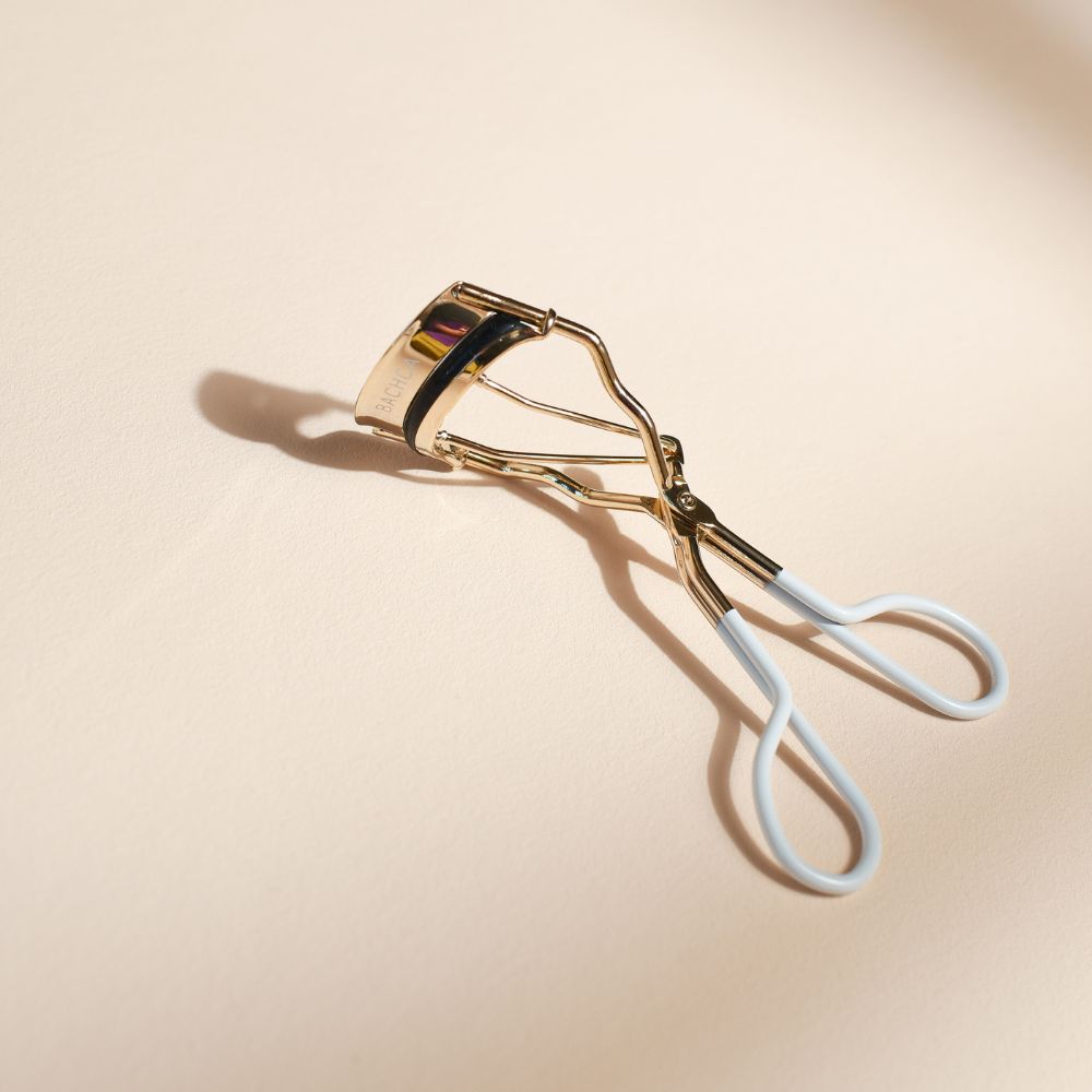 Recourbe-cils doré BACHCA avec poignées blanches en silicone, accessoire de maquillage élégant posé sur fond beige