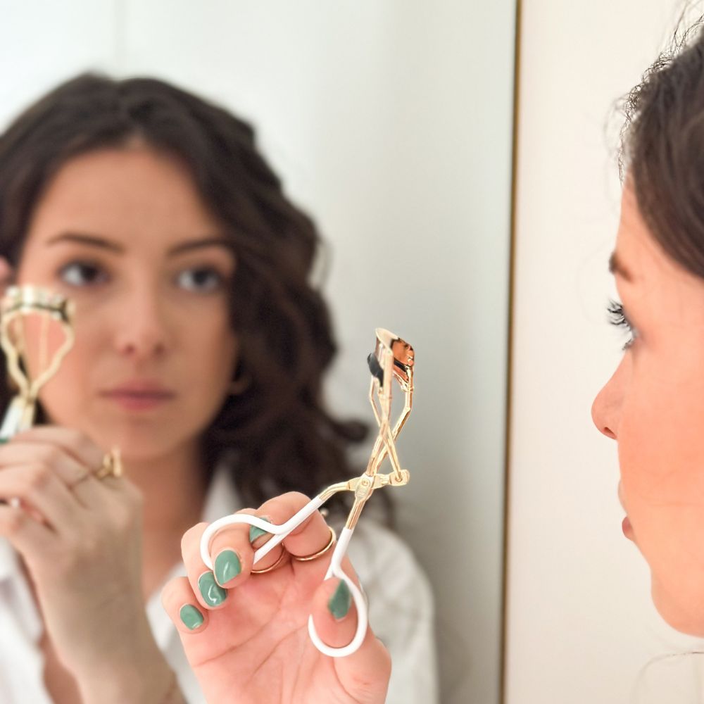 Femme utilisant le recourbe-cils doré Bachca devant un miroir, accessoire beauté élégant.